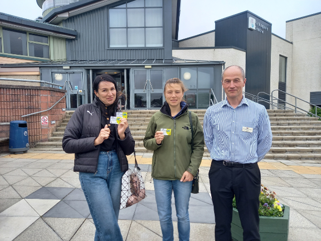 2 women standing in front of the Pickaquoy Centre with a man standing beside them holding up active life cards
