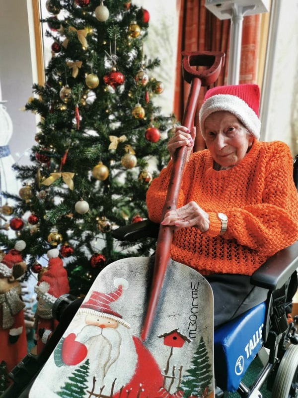 an elderly lady sitting beside a Christmas tree with a Santa hat on