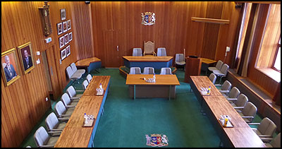a room with wooden walls, green carpet and tables and chairs in a horseshow shape with a table on the stage with 3 chairs