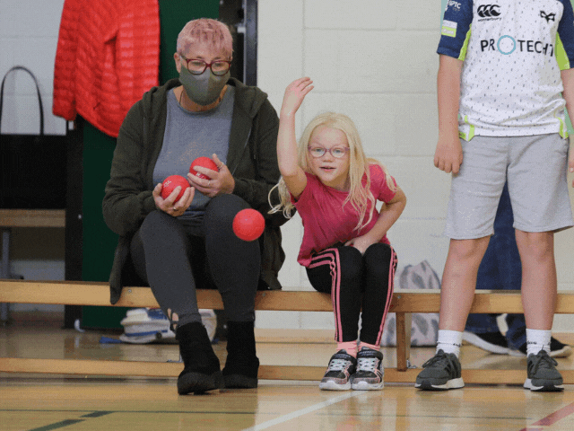 a lady watching a young child throw a ball whilst sitting on a bench