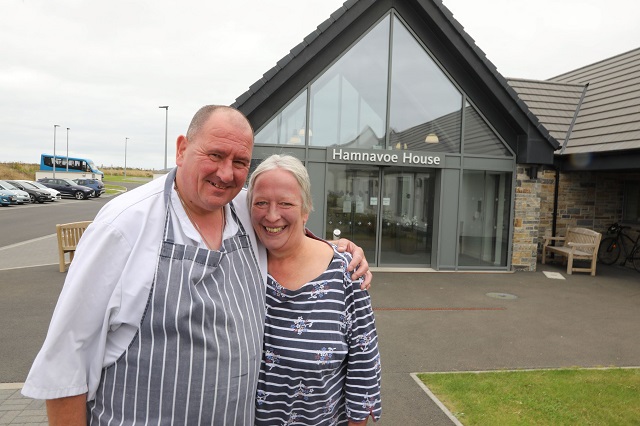2 people standing with their arms round each other in front of a building
