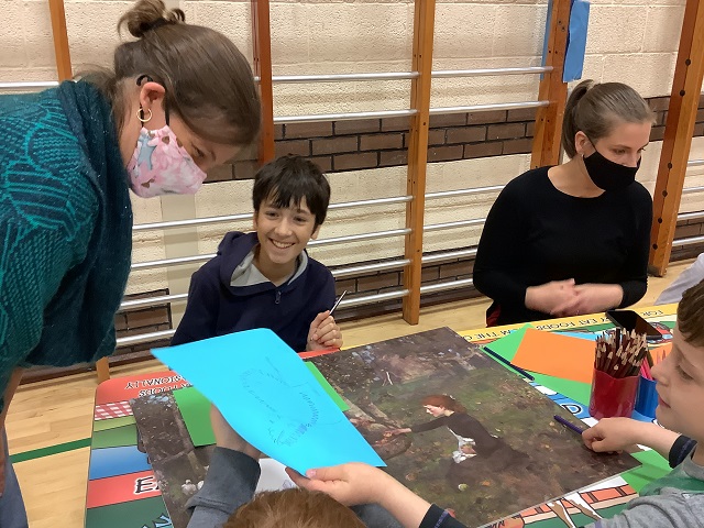 a lady standing over 2 children with a face mask looking at their artwork