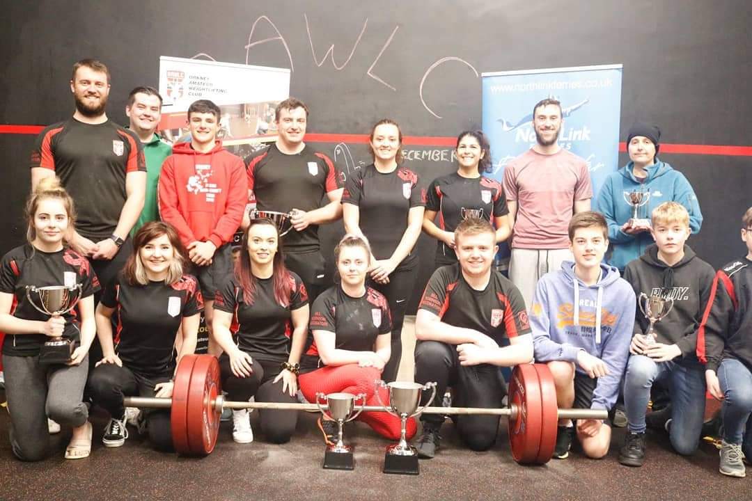 men, women and children gathering round a weight with trophies at the front