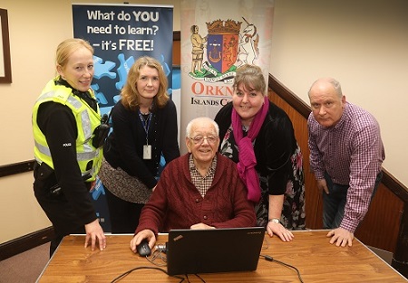 4 people standing over another person, one of these is a police officer,  huddled round a laptop showing him what to do