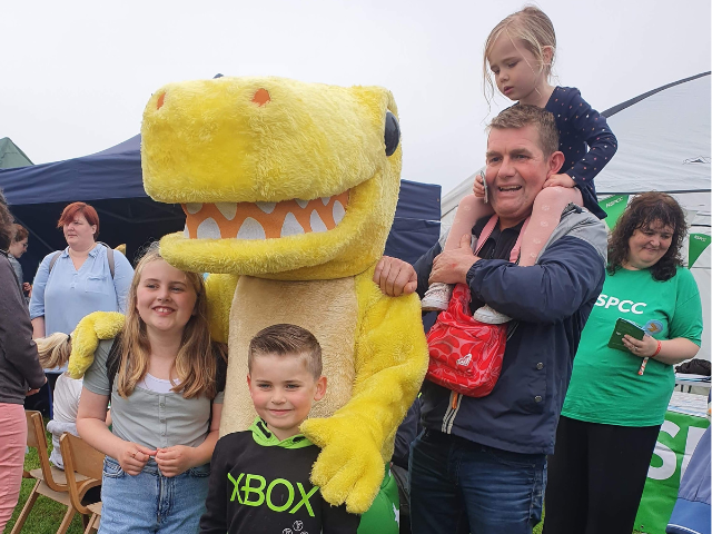 a yellow dinosaur hugging 2 children then a man beside them with a little girl on his shoulders
