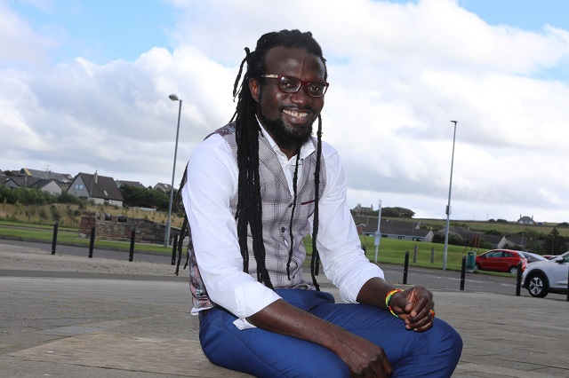 a man sitting with blue jeans and a white shirt with a grey waistcoat